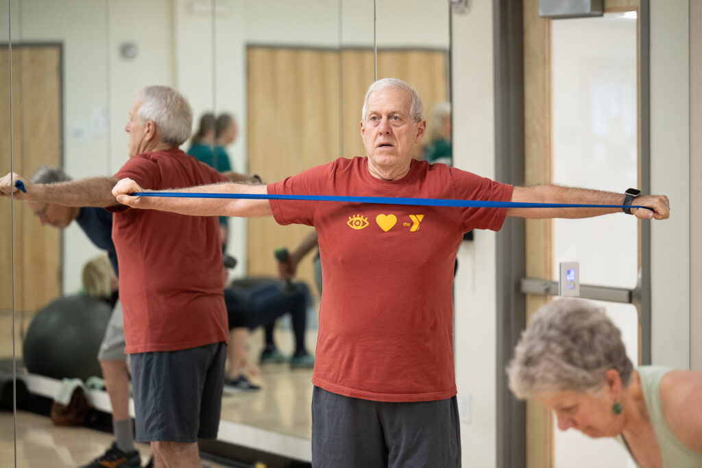 GBYMCA Active Older Adult fitness class: Man stretching long band, wearing an I LOVE THE Y shirt