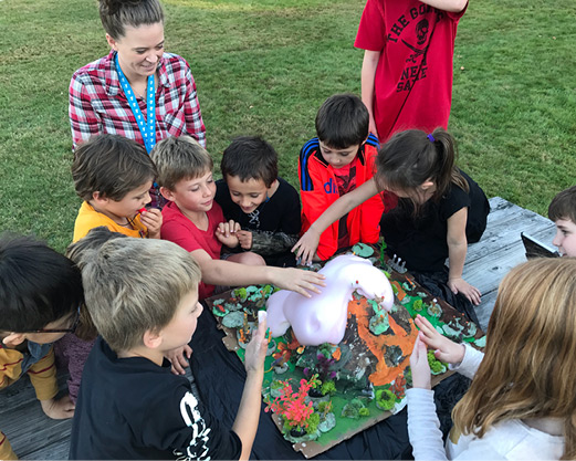 Youth outdoors gathered around a model volcano with pink lava