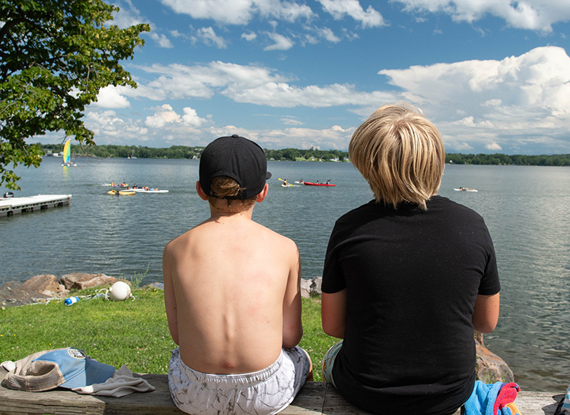 Two boys at GBYMCA Camp Abnaki with backs to viewer, looking out over the lake with kayakers