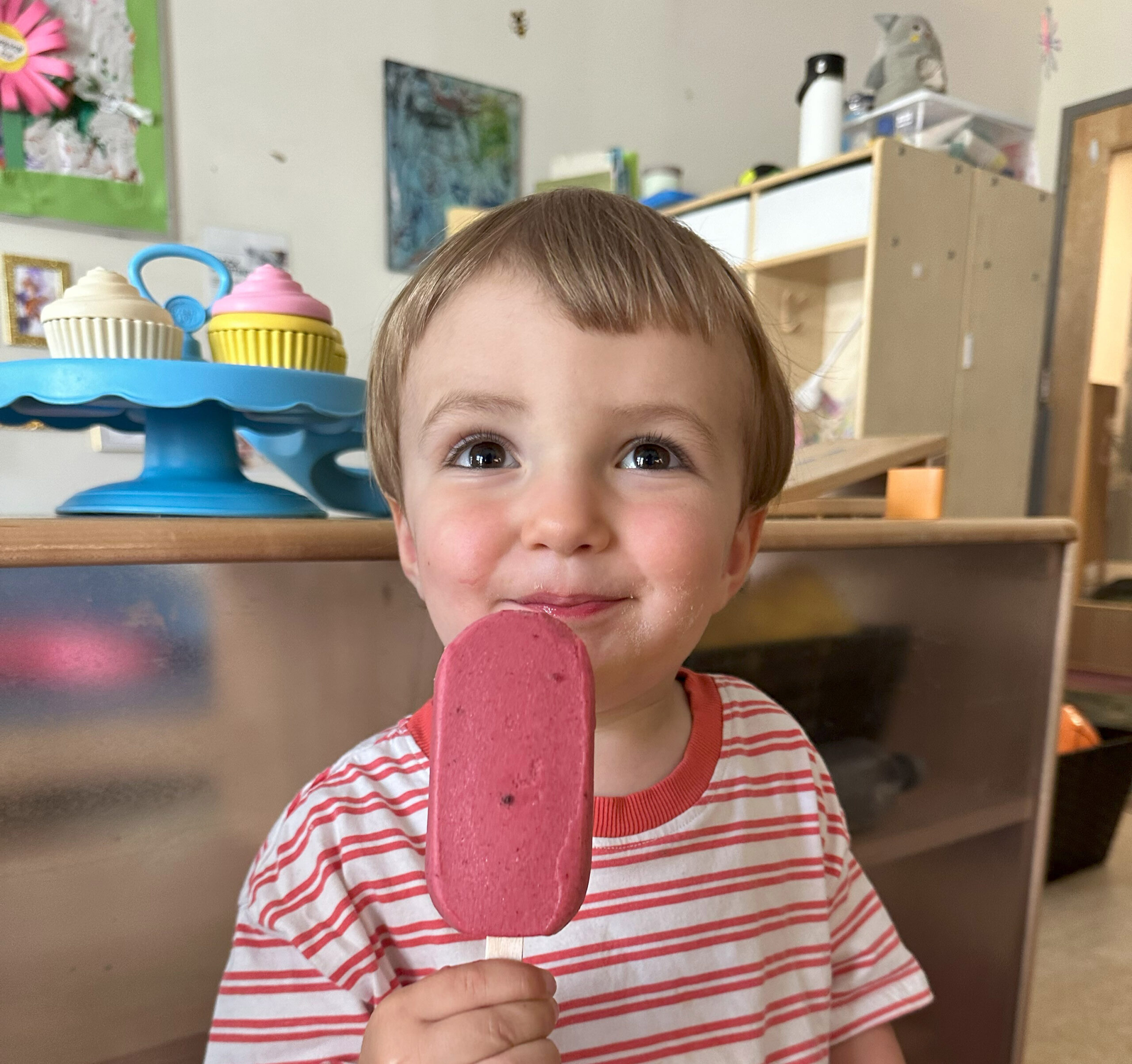 A young boy in a red and white striped shirt eats a red popsicle