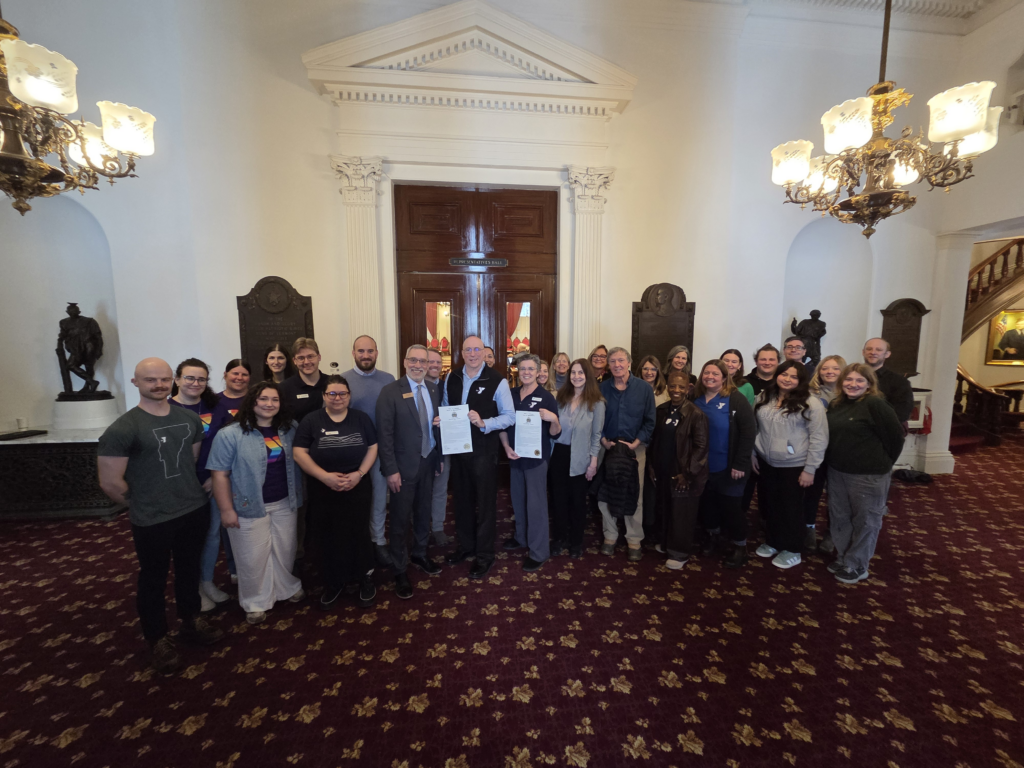 A crowd of people from the Y and Representatives from the VT Legislature are lined up in the lobby of the House floor with two copies of the 175th anniversary celebration Concurrent Resolution held by people in the middle