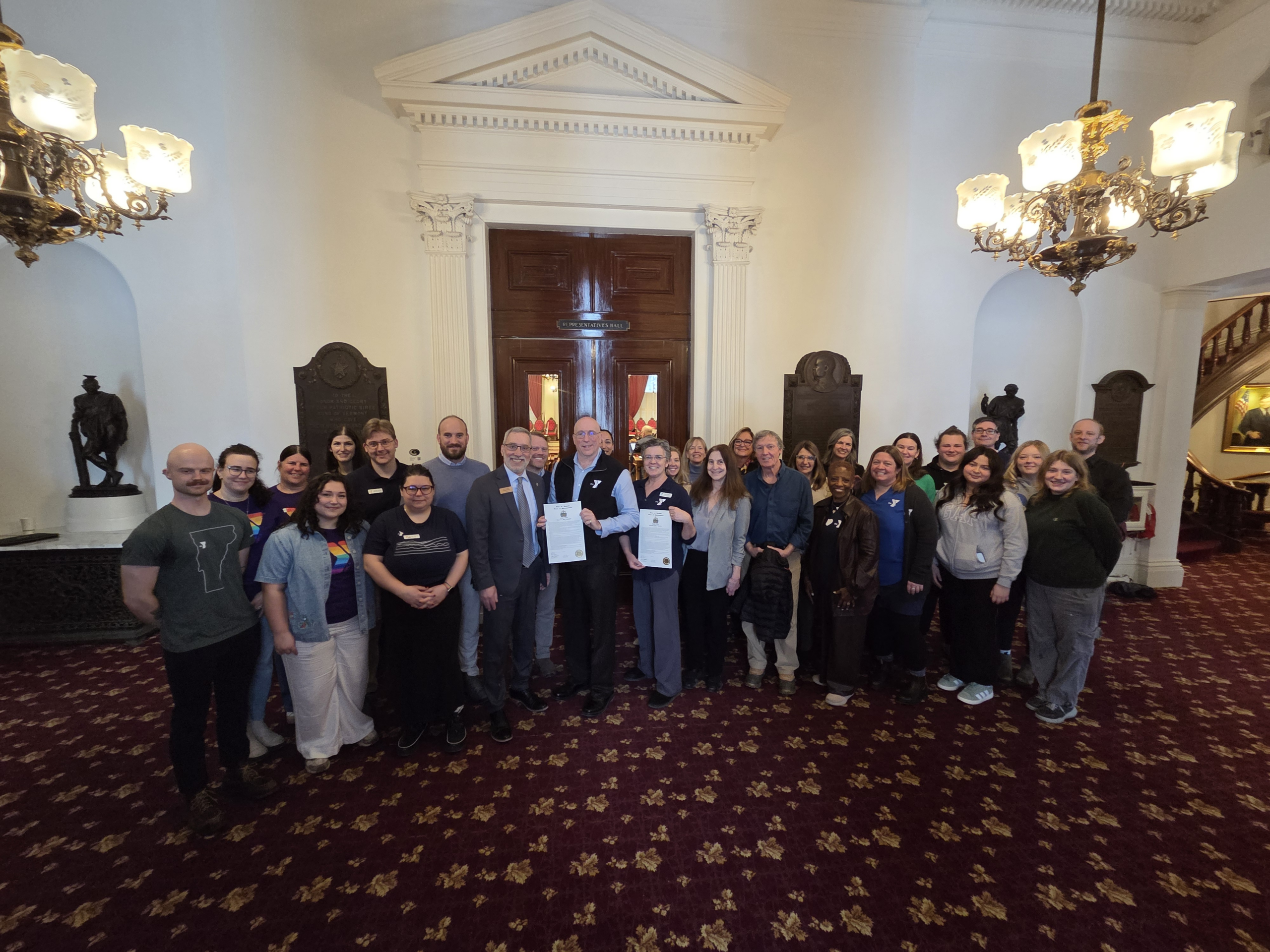 A crowd of people from the Y and Representatives from the VT Legislature are lined up in the lobby of the House floor with two copies of the 175th anniversary celebration Concurrent Resolution held by people in the middle