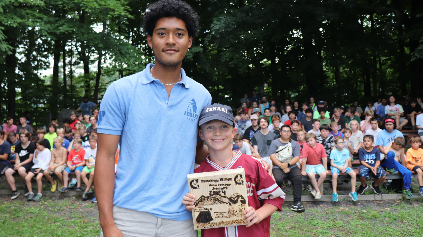 A camper and counselor stand in front of a crowd of summer camp boys seated behind them. The featured camper is holding his honor camper plaque.