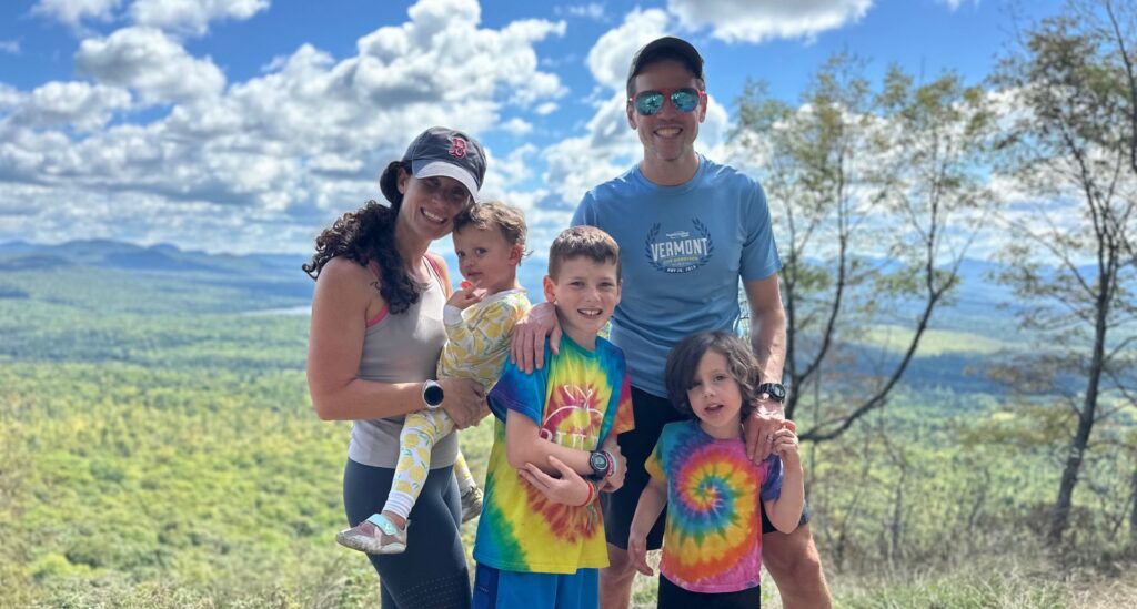 A family of five stands at the top of a mountain with blue sky and white puffy clouds